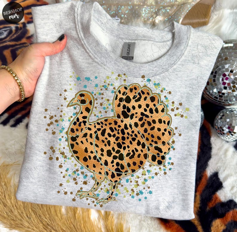 May include: A light gray sweatshirt featuring a turkey design with a leopard print pattern. The turkey is outlined in black and has brown and black spots. The design includes gold, blue, and brown speckles.