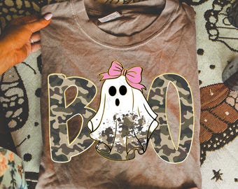 May include: A brown t-shirt with a graphic that says "BOO" in camouflage print. The "O" is a ghost wearing a pink bow. The shirt is folded and held by hands.