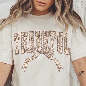 May include: Cream-colored t-shirt featuring the word "THANKFUL" in a floral design. A decorative bow with a floral pattern is below the word. The shirt has a relaxed fit and short sleeves, perfect for casual wear.