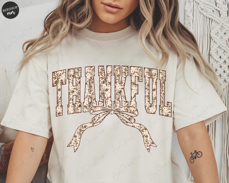 May include: Cream-colored t-shirt featuring the word "THANKFUL" in a floral design. A decorative bow with a floral pattern is below the word. The shirt has a relaxed fit and short sleeves, perfect for casual wear.