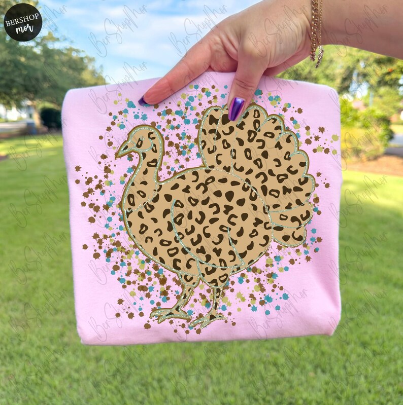 May include: A light pink t-shirt featuring a turkey design with a leopard print pattern. The turkey is outlined in gold and surrounded by brown and teal speckles. The shirt is held up by a person.