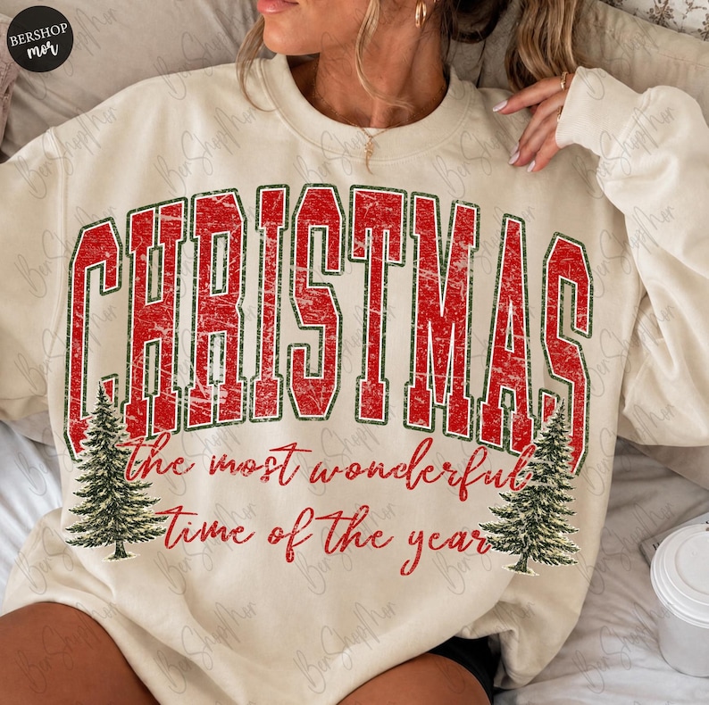 Op de afbeelding: Een cr&egrave;mekleurig sweatshirt met ronde hals met de rode, verweerde tekst CHRISTMAS in een groot collegiaal lettertype, met daaronder de rode cursieve tekst the most wonderful time of the year, geflankeerd door twee groene dennenboomillustraties.