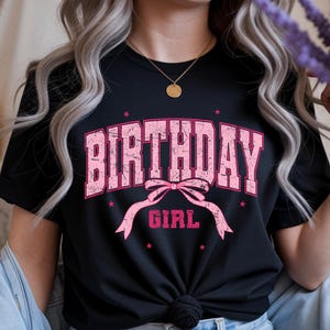 May include: Black t-shirt with the words "BIRTHDAY GIRL" in distressed pink lettering. The design includes a pink bow and stars. The shirt is a casual style.