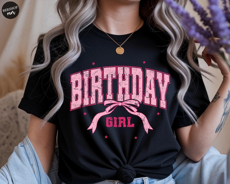 May include: Black t-shirt with the words "BIRTHDAY GIRL" in distressed pink lettering. The design includes a pink bow and stars. The shirt is a casual style.