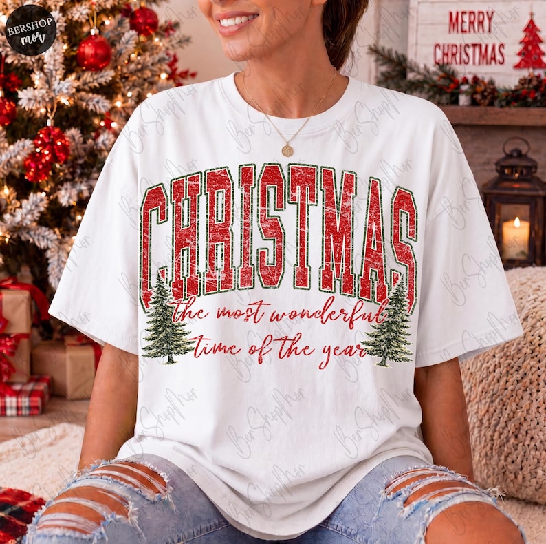 Op de afbeelding: Een wit t-shirt met korte mouwen voorzien van de rode verweerde tekst CHRISTMAS met groene omlijningen, en de rode cursieve tekst the most wonderful time of the year, vergezeld door twee groene dennenboomillustraties aan de zijkanten.