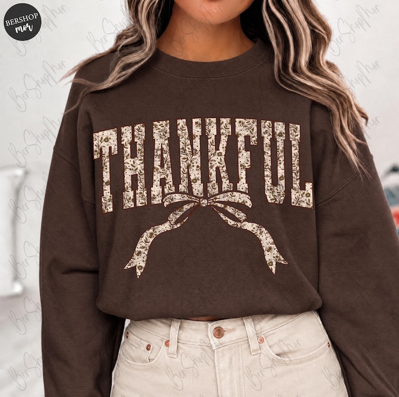 May include: A brown sweatshirt with the word "THANKFUL" in a floral design across the chest. A decorative bow is below the word. The sweatshirt is a solid brown color.