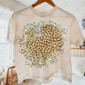 May include: Cream-colored t-shirt featuring a turkey design with a leopard print pattern. The turkey is surrounded by teal, brown, and green paint splatters. The shirt is hanging on a wooden hanger.