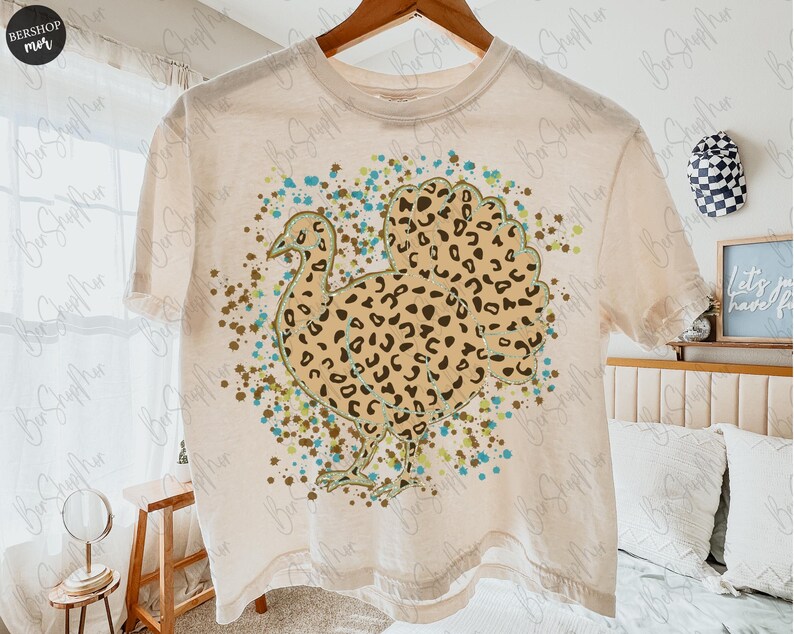 May include: Cream-colored t-shirt featuring a turkey design with a leopard print pattern. The turkey is surrounded by teal, brown, and green paint splatters. The shirt is hanging on a wooden hanger.