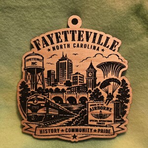 May include: A wooden ornament featuring a laser-engraved skyline of Fayetteville, North Carolina. The design includes a water tower, a train, a parachutist, and text reading FAYETTEVILLE NORTH CAROLINA, HOME OF THE AIRBORNE & SPECIAL OPERATIONS FORCES, and HISTORY COMMUNITY PRIDE.