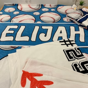 Op de afbeelding: Een blauwe fleece deken met een baseballthema. De deken heeft witte baseballs met rode naden en de naam "Elijah" gedrukt in witte letters. Een witte fleece deken met zwarte tekst en een rood ontwerp is gevouwen op de blauwe deken.