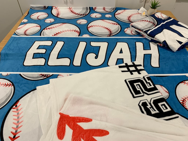 Op de afbeelding: Een blauwe fleece deken met een baseballthema. De deken heeft witte baseballs met rode naden en de naam "Elijah" gedrukt in witte letters. Een witte fleece deken met zwarte tekst en een rood ontwerp is gevouwen op de blauwe deken.