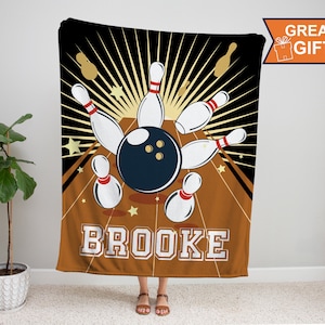 May include: A brown and orange fleece blanket with a black and white illustration of bowling pins and a bowling ball. The text "BROOKE" is printed in white letters on the blanket.