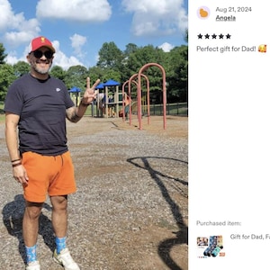 Pode incluir: Uma pessoa vestindo uma camiseta preta, shorts laranja, bon&eacute; vermelho e branco e &oacute;culos de sol, fazendo o sinal de paz em um parque. O c&eacute;u &eacute; azul com nuvens brancas. O texto diz "Perfect gift for Dad!"