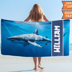 May include: A blue beach towel with a white shark swimming in the ocean. The towel has the name "William" printed on it in white letters.