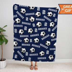 May include: A navy blue fleece blanket with a repeating pattern of white soccer balls and the name "Brooke" and the number "2" in white.