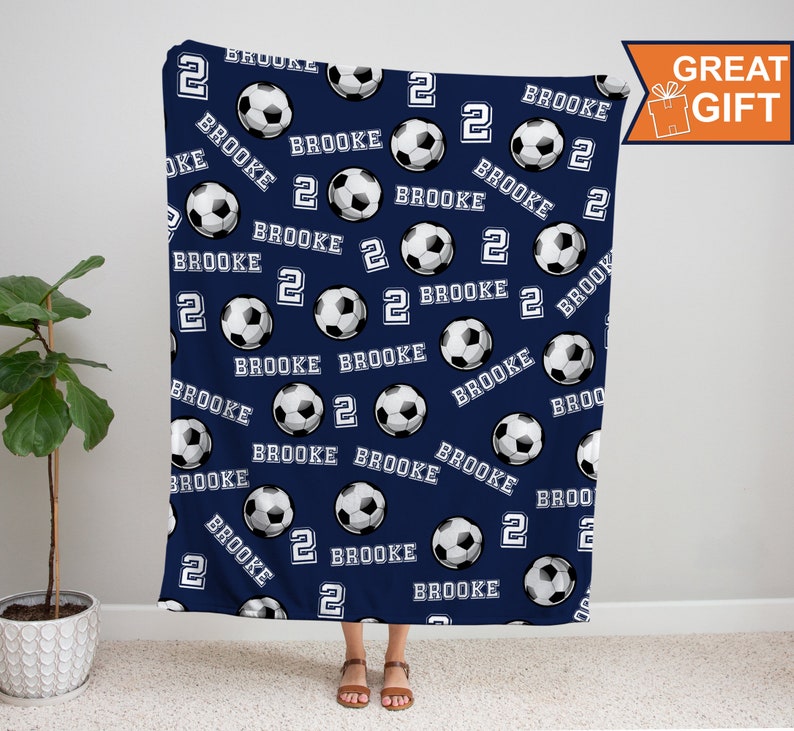 May include: A navy blue fleece blanket with a repeating pattern of white soccer balls and the name "Brooke" and the number "2" in white.