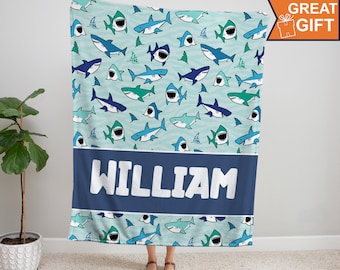 May include: A blue and green fleece blanket with a pattern of cartoon sharks. The blanket has a navy blue band with the name "WILLIAM" printed in white.