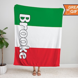 May include: A green, white, and red fleece blanket with the name "Brooke" printed in white on the left side. The blanket is folded in half and standing upright.