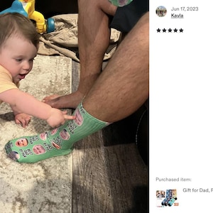 Pode incluir: Uma meia verde clara com fotos e texto personalizados, com um beb&ecirc; apontando para ela. A meia tem v&aacute;rias imagens e o texto "BEST DAD BY FAR". Um beb&ecirc; com uma camisa amarela est&aacute; engatinhando em um tapete.