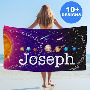 May include: A purple beach towel with a colorful solar system design and the name "Joseph" printed in white. The towel is held by a person in front of a beach scene.