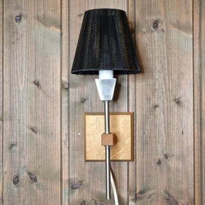 Peut inclure: Une lampe murale avec un abat-jour noir et une tige argentée. La lampe est fixée à une base carrée en bois avec un petit bloc de bois. La lampe est fixée sur un mur en bois.