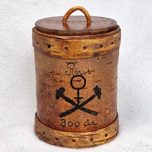 Puede incluir: Un recipiente de madera con tapa, hecho de corteza de abedul. Tiene un diseño en blanco y negro de un martillo y un símbolo, con el texto "Roros 300 år" en la parte delantera.