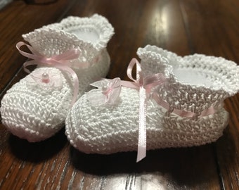 May include: A pair of white crocheted booties featuring a scalloped ankle cuff, a delicate light pink satin ribbon threaded through the top, and a small decorative light pink fabric flower with a central white bead on the front of each shoe.