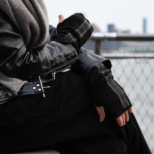 May include: A person wearing a black leather jacket with a gray scarf and black fingerless gloves. The person is sitting on a concrete surface.