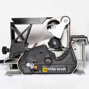 May include: A close-up of a Work Sharp Knife Sharpener with a silver and black body. The sharpener has a belt system and a tracking adjustment knob. The text "Work Sharp Sharpeners" is printed on the side.