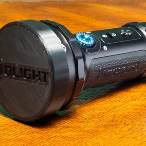 May include: Black flashlight with a blue button and the word "OLIGHT" on the end cap. The flashlight has a textured grip and several buttons and switches. The flashlight is on a wooden surface.