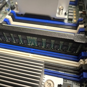 May include: A close-up of a computer motherboard with two black RAM sticks installed in the slots. The RAM sticks have blue clips on either side and are partially inserted into the slots.