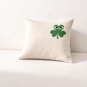 May include: Square cream-colored pillow with a green shamrock design. The shamrock has a face with black eyes. The pillow is made of a soft, textured fabric, perfect for home decor. The design is embroidered.