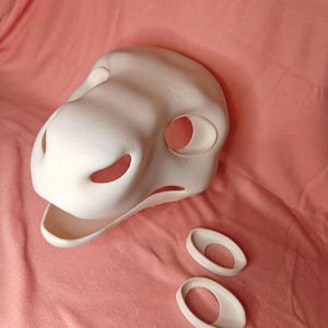 May include: A white, unpainted fursuit head with large eye openings and a wide mouth. Two matching white eye blanks are positioned below the head. The mask is designed to be customized and painted.