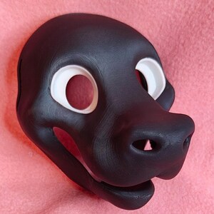 Op de afbeelding: Een zwart dierenmasker met witte oogomrandingen. Het masker heeft een prominente snuit met twee neusgaten en een lachende mond. Waarschijnlijk voor cosplay of kostuums.