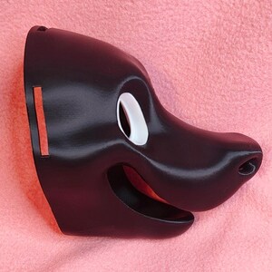Op de afbeelding: Een zwart masker in de vorm van een cartoon-hondengezicht. Het masker heeft een wit oog, een lachende mond en een prominente neus. Het masker is gemaakt van een glad, glanzend materiaal.