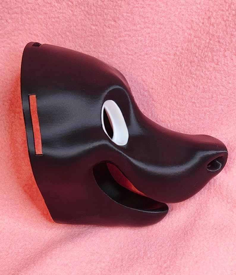 Op de afbeelding: Een zwart masker in de vorm van een cartoon-hondengezicht. Het masker heeft een wit oog, een lachende mond en een prominente neus. Het masker is gemaakt van een glad, glanzend materiaal.