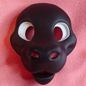 Op de afbeelding: Een zwart dierenmasker met grote, witte oogopeningen en een prominente snuit. Het masker is gemaakt van een glad materiaal en heeft een gedetailleerd ontwerp, inclusief neusgaten en een mond. Het masker is ontworpen om gedragen te worden.