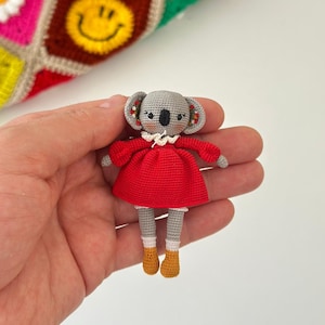 May include: A handmade crochet koala doll in a red dress, white collar, and yellow boots. The doll has grey fur and detailed features. A colourful crochet blanket with daisy and smiley face squares is in the background.