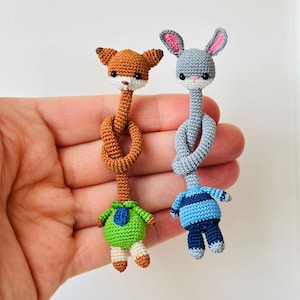 May include: Two handmade crochet animal toys: a fox and a bunny. The fox is brown with a white face and a green outfit. The bunny is gray with pink ears and a blue outfit. Both toys have long necks knotted in the middle.