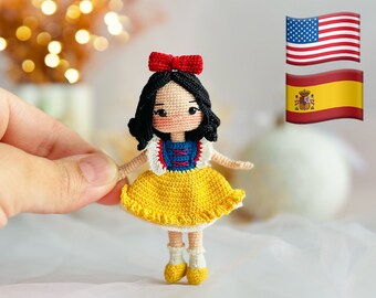 May include: A crocheted doll of Snow White wearing a yellow dress with a blue bodice and red bow. The doll is standing on a white surface with a blurred background of lights. The American and Spanish flags are visible in the background.