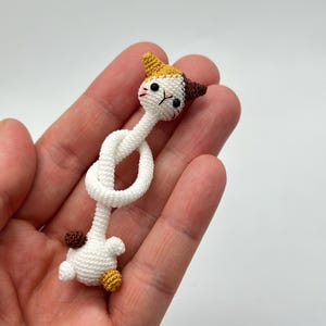 May include: A handmade crochet cat figurine in white, brown, and yellow. The cat has a long, knotted body and a detailed face with black eyes and whiskers. The ears are yellow and brown. The cat's body is white.