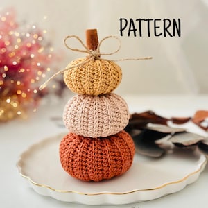 May include: A stack of three crocheted pumpkins in shades of orange, peach, and yellow, topped with a cinnamon stick and twine bow. The pumpkins sit on a white plate with a gold rim. The word "PATTERN" is written in black.