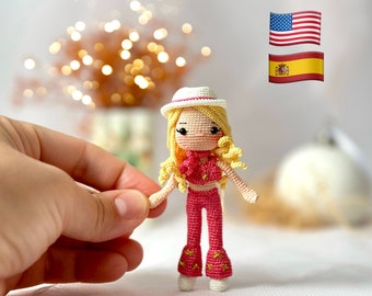 May include: A crocheted doll with blonde hair, wearing a white hat, pink top, and pink pants. The doll is standing on a white surface with a blurred background.