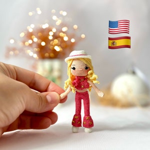 Peut inclure: Une poupée en crochet aux cheveux blonds, portant un chapeau blanc, un haut rose et un pantalon rose. La poupée est debout sur une surface blanche avec un arrière-plan flou.