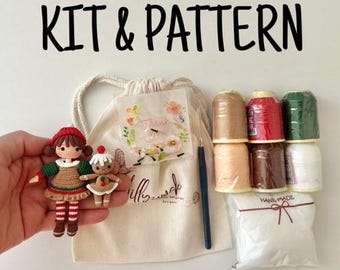 以下が含まれることがあります： 「KIT & PATTERN」の文字が入ったクラフトキット。キットには人形、巾着袋、糸巻き、その他のクラフト用品が含まれています。人形は赤い帽子と緑と赤の衣装を着ています。バッグには花柄のデザインがあります。