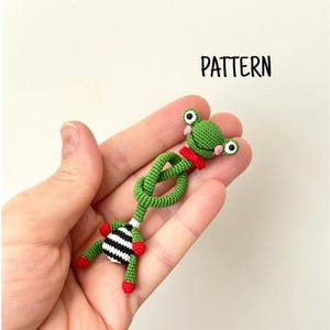 May include: A handmade green crochet frog with red details and a black and white striped belly. The frog has large eyes and is held in a hand. The word "PATTERN" is in the background.