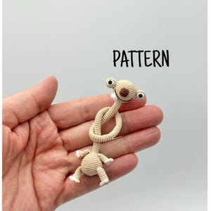 Puede incluir: Una muñeca de ganchillo beige hecha a mano con un cuerpo anudado y un diseño caprichoso. La muñeca tiene ojos grandes y redondos, una nariz pequeña y una boca sonriente. La palabra "PATTERN" está escrita en texto negro.