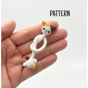 Puede incluir: Un juguete para gatos tejido a ganchillo en blanco, crema y marrón con un cuerpo anudado. La cabeza y las patas del gato están detalladas con detalles marrones y amarillos. La palabra "PATTERN" está impresa en negro.