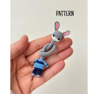 Może przedstawiać: Szydełkowa zabawka króliczek i lalka z szarymi i niebieskimi detalami. Króliczek ma długie uszy i białą twarz. Lalka ma na sobie niebiesko-szarą koszulę w paski i niebieskie spodnie. Słowo "PATTERN" znajduje się w prawym górnym rogu.