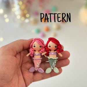 May include: Two small crocheted mermaid dolls with the word PATTERN printed above them. One doll has pink hair and a pink tail, while the other has red hair and a green tail. Both dolls feature light skin tones, embroidered facial details, and small tops.